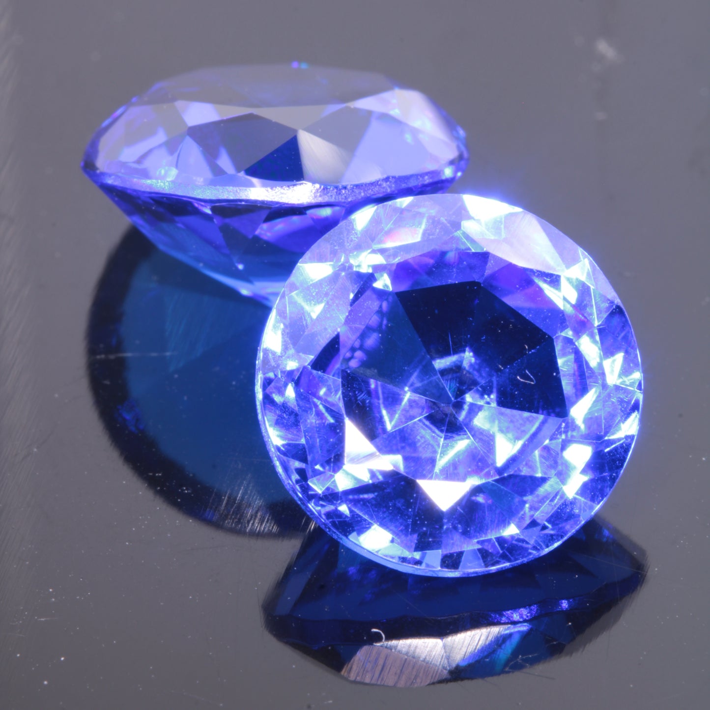 Two Faceted Round Djeva Blue Lab Spinel Gemstones, 12 mm in diameter on a dark background.