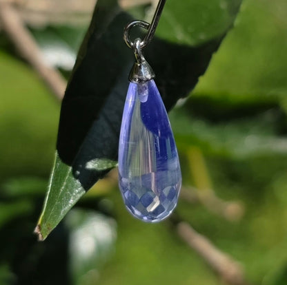 A faceted teardrop-shaped fluorite droplet earring with a blue fluorescent glow color, hanging against a blurred green leaf background.