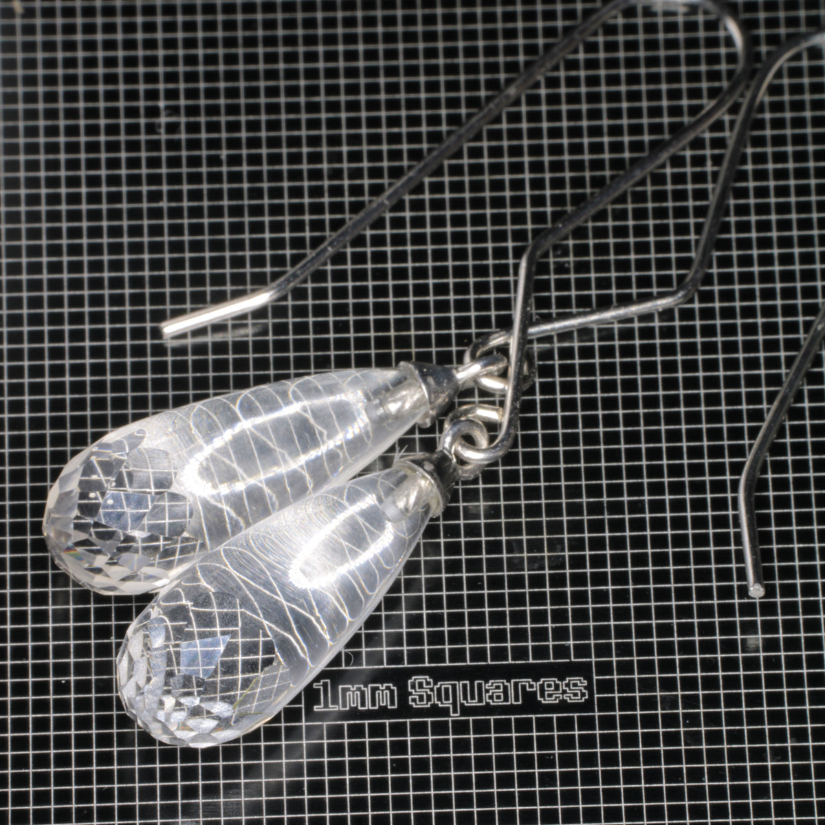 Clear faceted teardrop-shaped fluorite earrings with sterling silver fishhook ear wires, displayed on a black grid mat labeled '1mm Squares'.