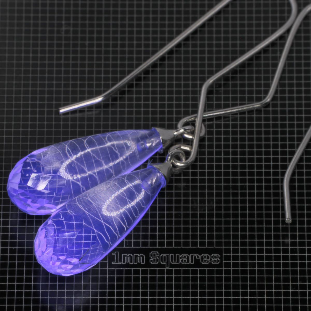 Clear faceted teardrop-shaped glowing blue fluorite earrings with sterling silver fishhook ear wires, displayed on a black grid mat labeled '1mm Squares'.
