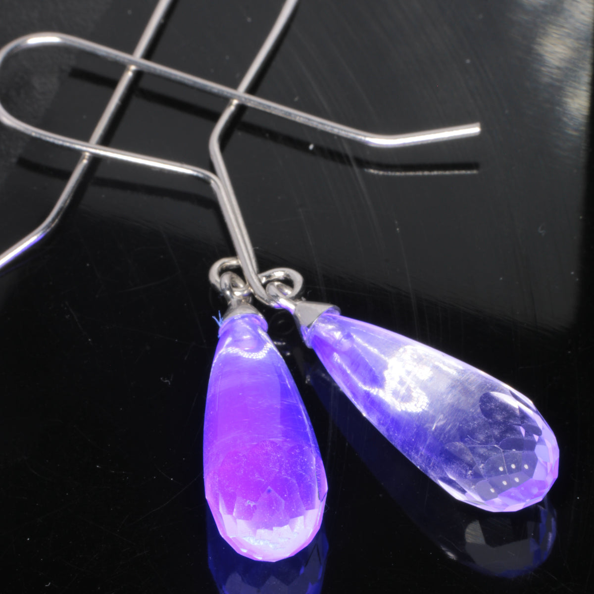 Clear faceted teardrop-shaped fluorite earrings with sterling silver fishhook ear wires, displayed on a black metal surface under UV showing a blue glow