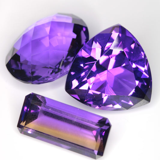Three Faceted Hydrothermal Amethyst Gemstones, grown in the Soviet Union and cut into a checkerboard oval, a trillion, and a long bar.  The long bar is striped yellow and purple due to citrine zones.